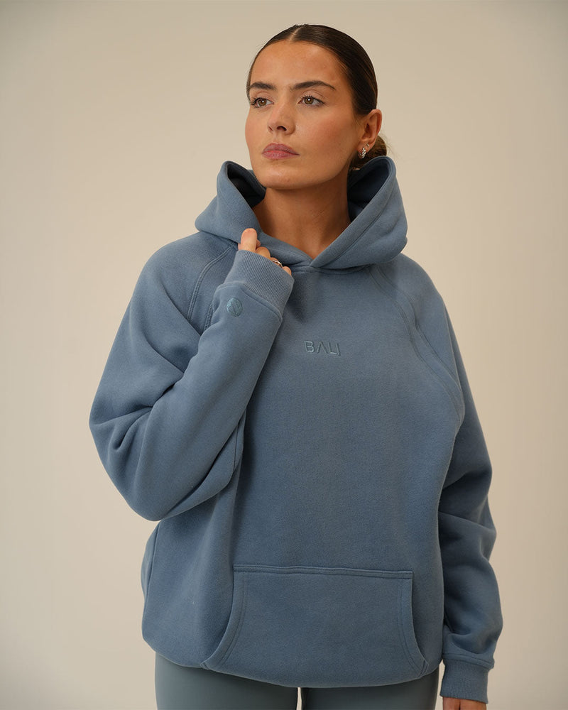 A woman wearing a dusty blue pullover BALI hoodie with a small tonal embroidered "BALI" logo on the chest, posed against a neutral beige background.