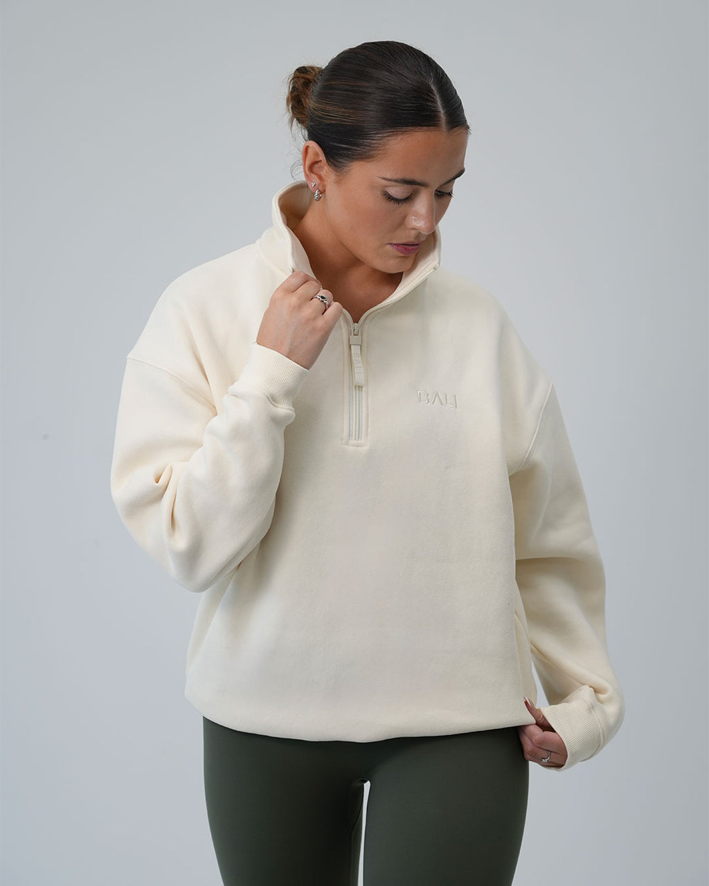 A woman wearing a Signature BALI oat-coloured half-zip sweatshirt and dark olive green leggings, standing against a neutral light grey studio background.
