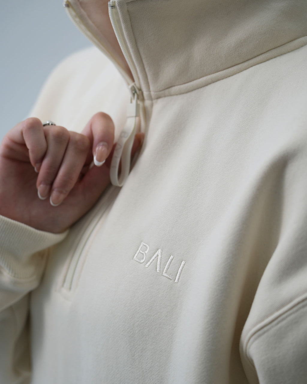 Close-up of a person wearing a Signature BALI oat-coloured half-zip sweatshirt with the brand name 'BALI' embroidered on the chest.