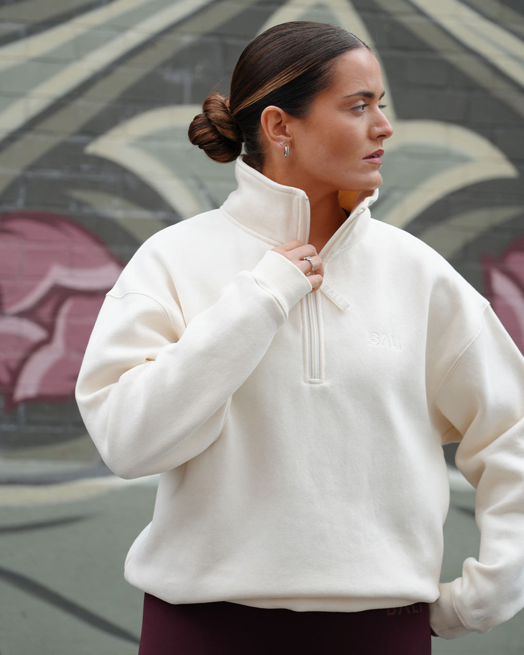 A woman wearing an oat BALI signature half-zip sweatshirt with a drawstring collar, standing in front of a graffiti-covered wall.