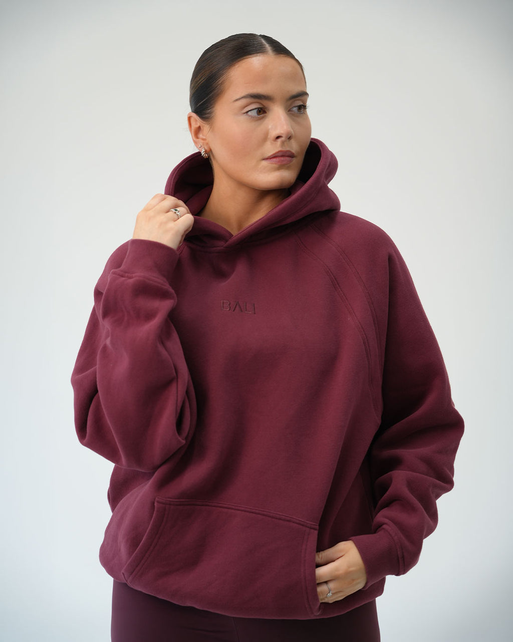 A woman wearing an oversized maroon Bali hoodie with a tonal embroidered logo "Bali" on the chest, posed against a light gray studio background.
