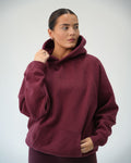 A woman wearing an oversized maroon Bali hoodie with a tonal embroidered logo "Bali" on the chest, posed against a light gray studio background.