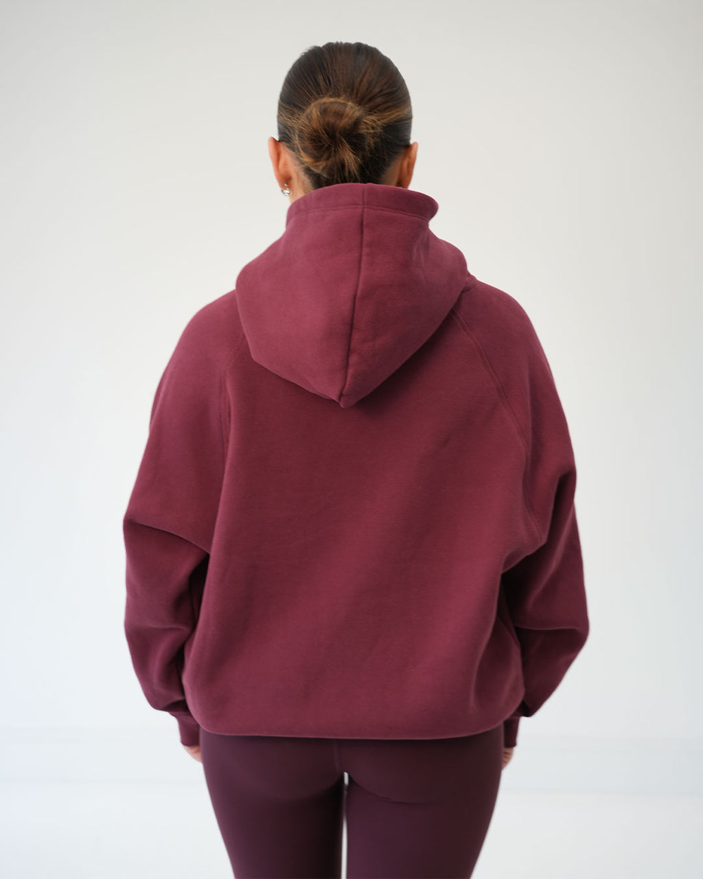 A Woman viewed from behind wearing a loose-fitting maroon hooded sweatshirt and matching leggings against a plain light gray studio background.