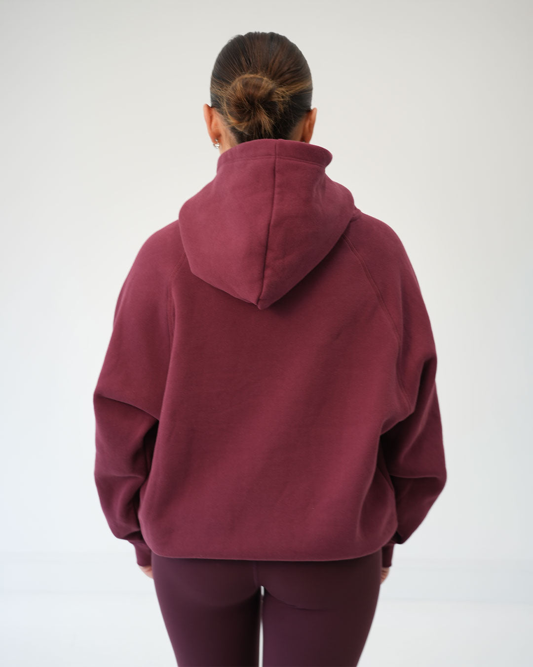 A Woman viewed from behind wearing a loose-fitting maroon hooded sweatshirt and matching leggings against a plain light gray studio background.