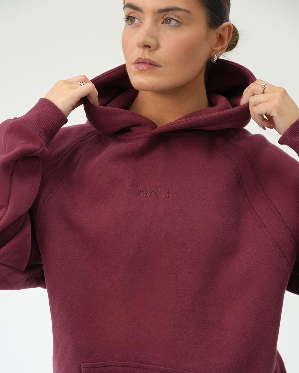 A woman wearing a maroon pullover hoodie with 'BALI' embroidered on the chest, posing with her hands on the hood against a light gray background.