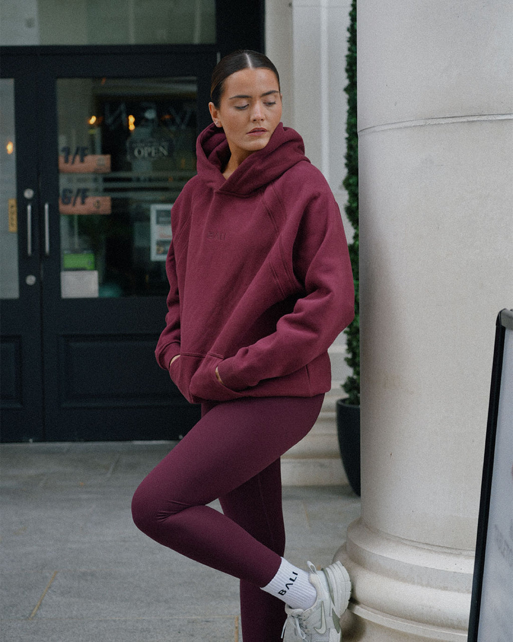 A woman wearing a matching BALI burgundy hoodie and leggings set, white socks, and white sneakers, posing outdoors next to a large stone column in front of a storefront.