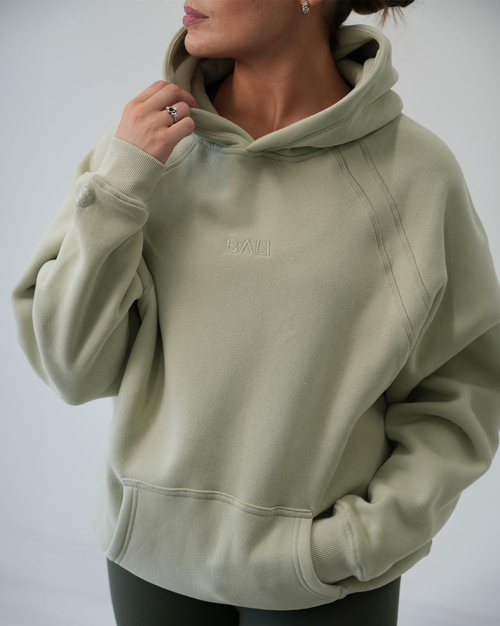 A woman wearing an oversized sage green BALI hoodie with a small embroidered 'BALI' logo on the chest, shown from the neck down against a light gray studio background.