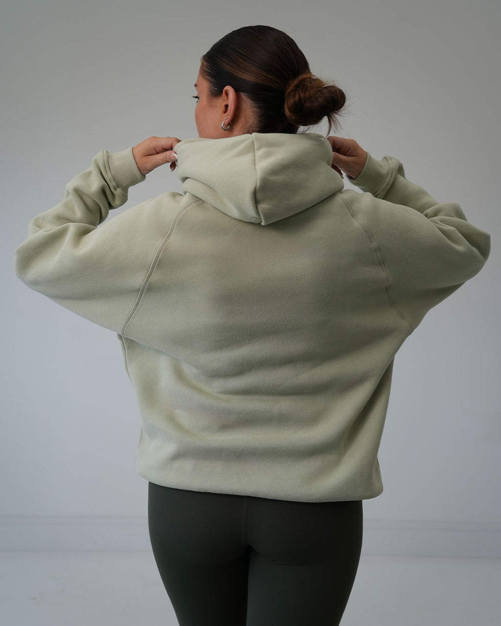 A woman viewed from behind wearing a BALI luxury sage green hoodie and dark leggings, adjusting the hood against a plain light gray studio background.