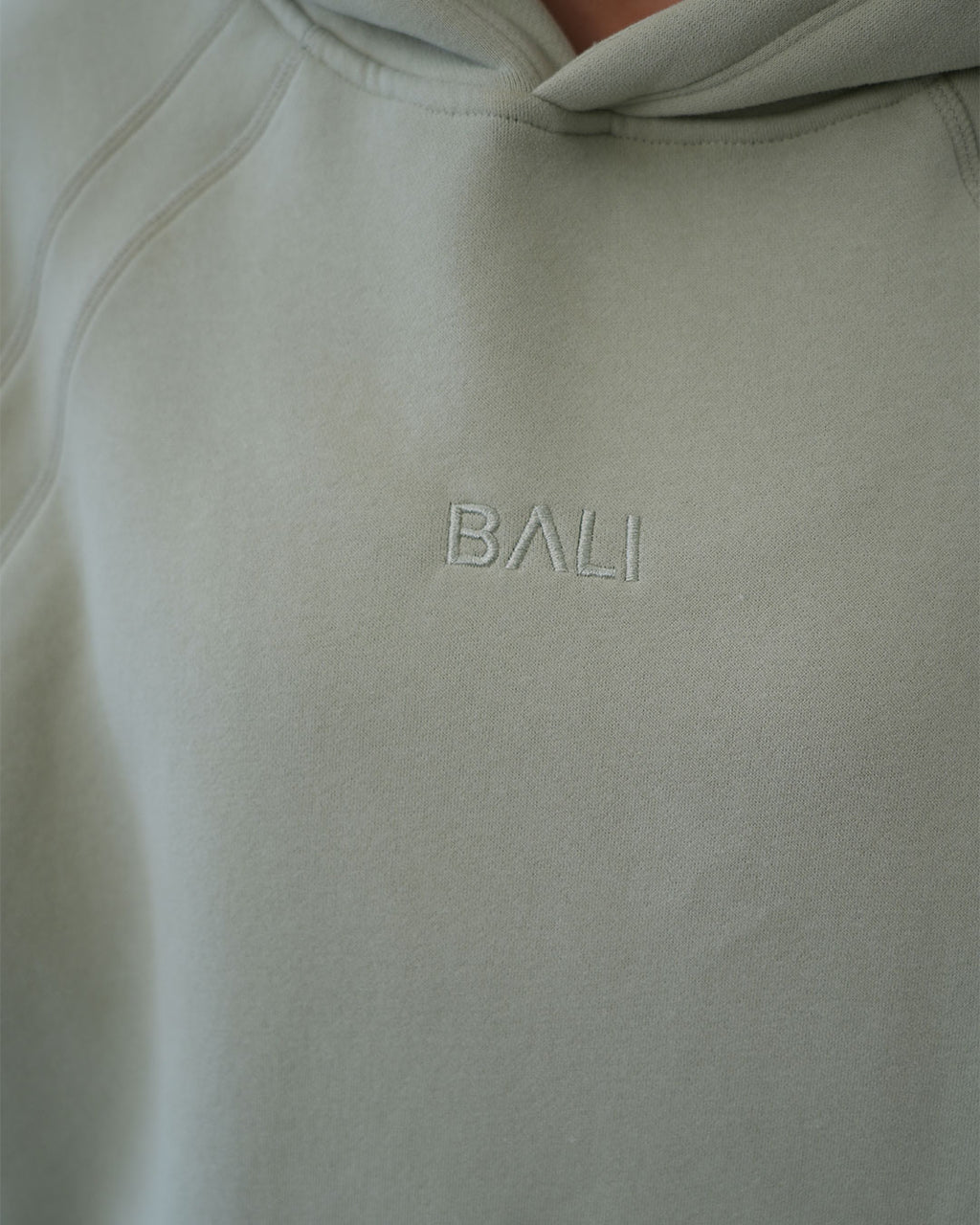 Close-up of a light sage green luxury hoodie with the word 'BALI' embroidered in matching thread on the chest.