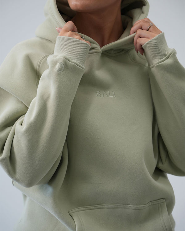 A close-up of a person wearing a light sage green luxury hoodie with the brand name 'BALI' subtly embroidered on the chest. The person is holding the hood up with both hands against a plain light gray background.