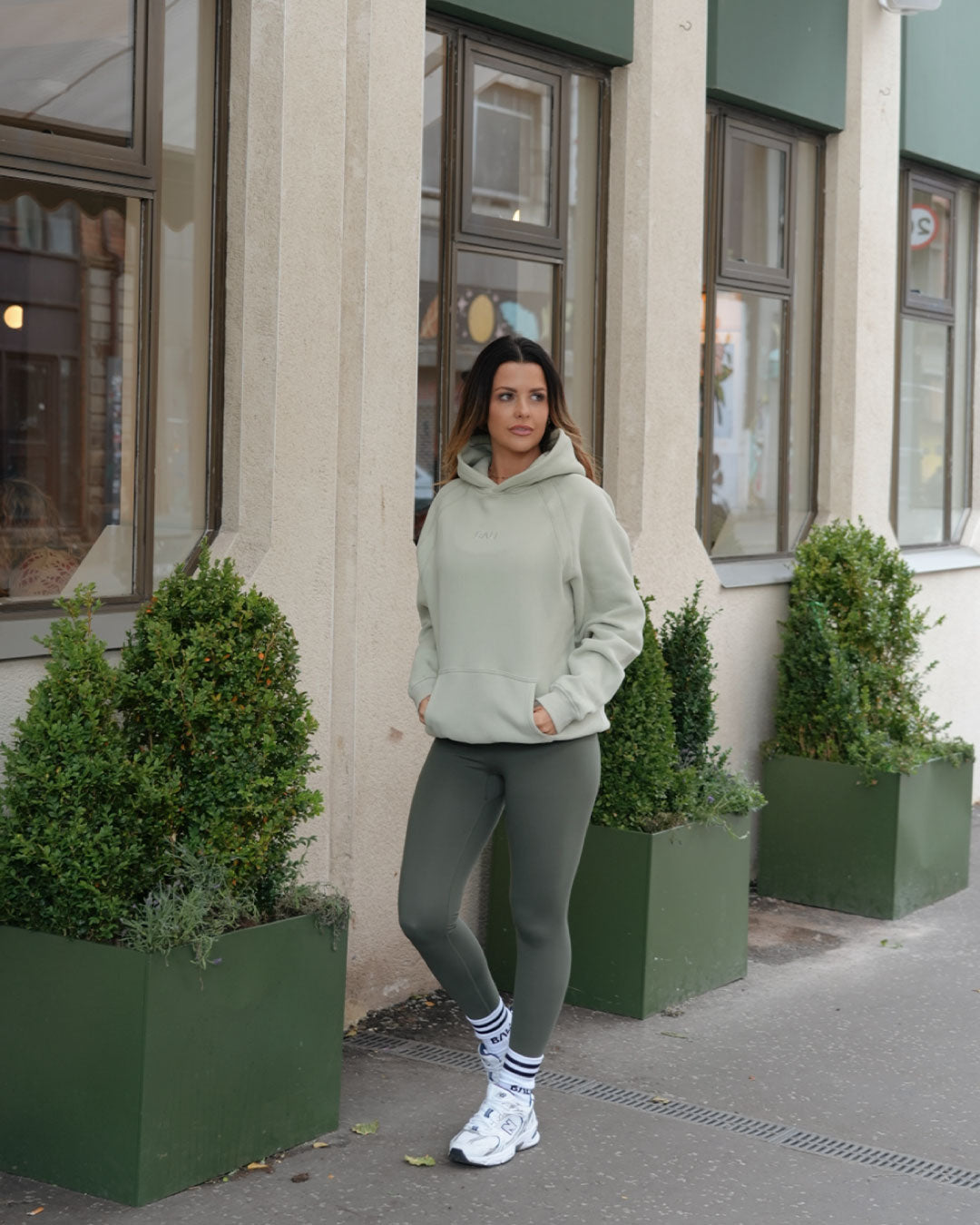 A woman stands outdoors in front of a building, wearing a light sage green  BALI hoodie, olive green BALI leggings, white socks, and white sneakers.