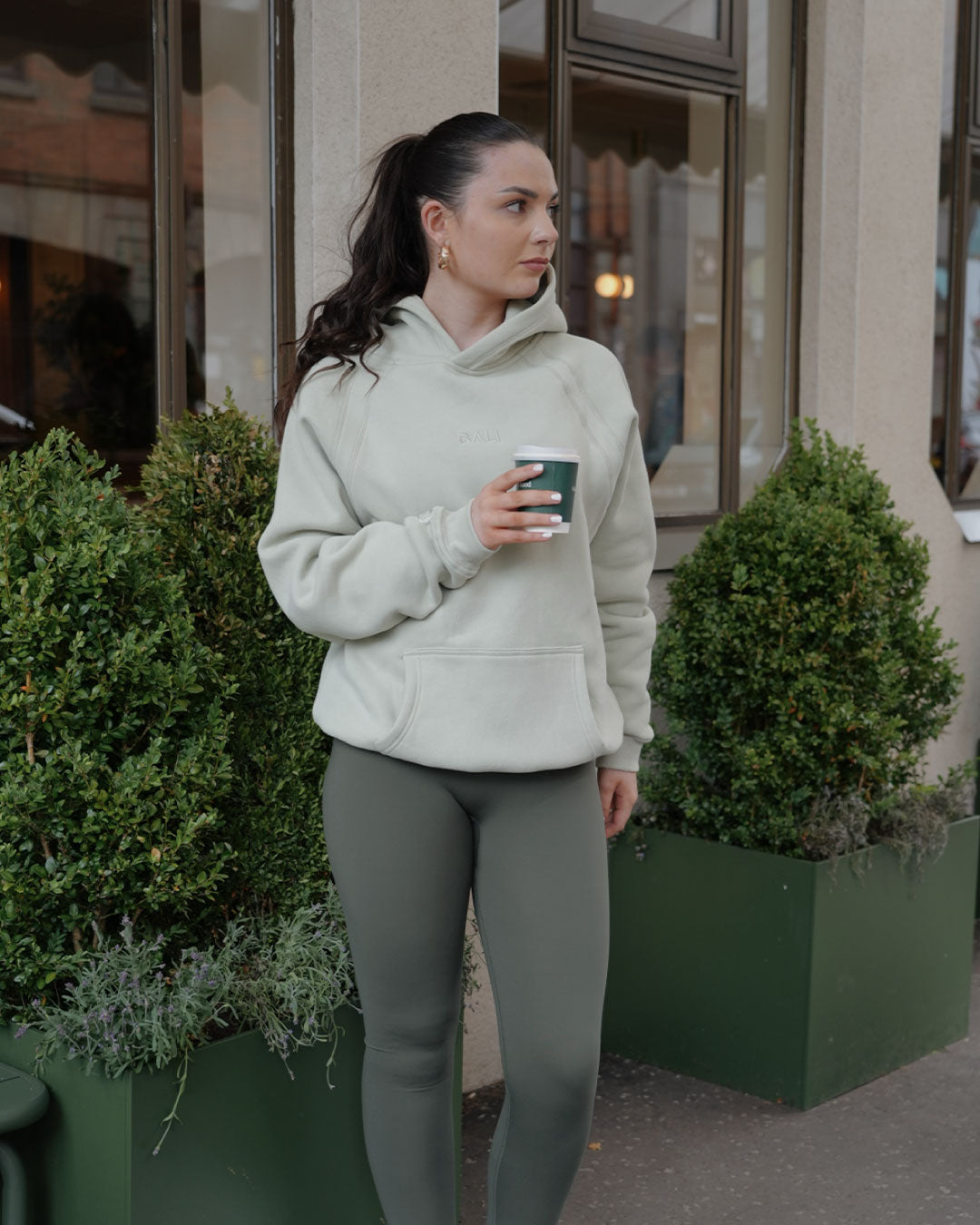 A woman wearing a light sage green BALI hoodie and matching sage green BALI leggings stands outdoors in front of a building, holding a dark green coffee cup.