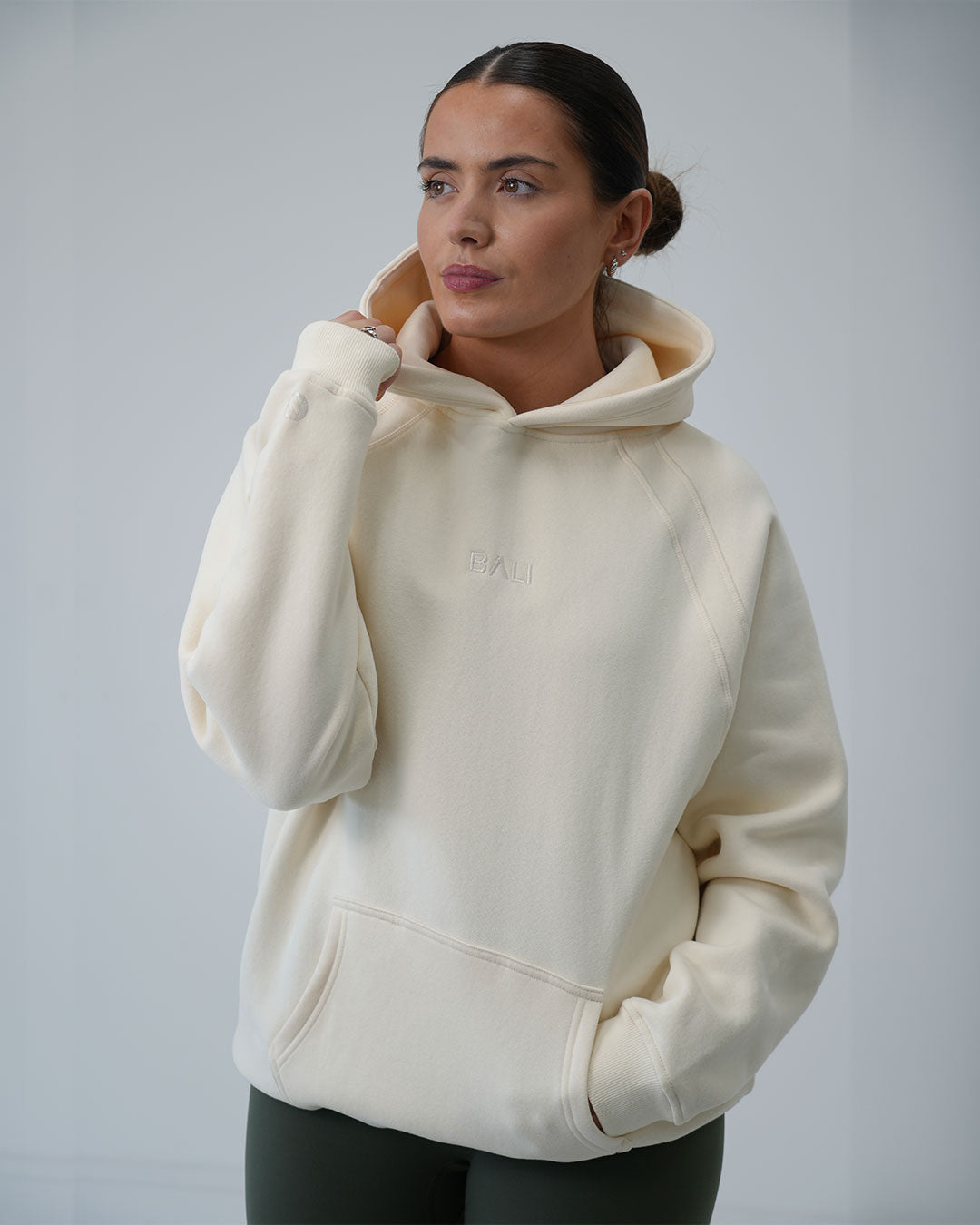 A woman wearing a cream-colored BALI oversized hoodie with a tonal embroidered "BALI" logo on the chest, posed against a neutral light gray background.