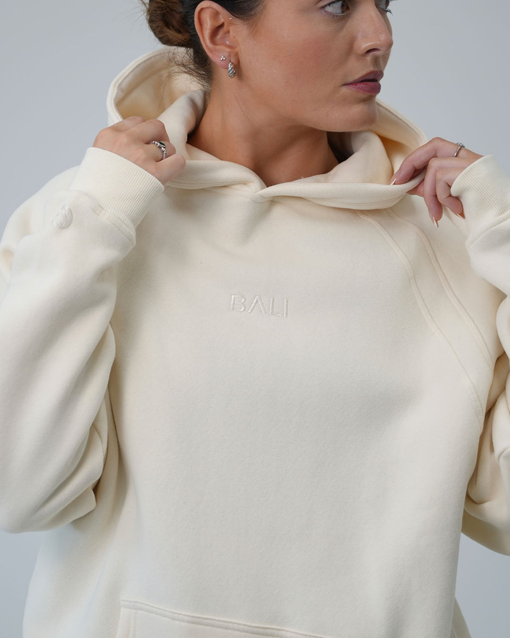 A close-up of a woman wearing a cream-colored BALI hoodie with the brand name 'BALI' subtly embroidered on the chest, set against a plain light gray background.