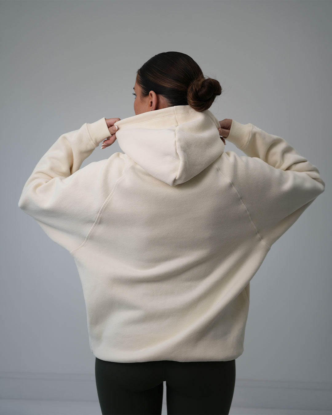 A woman viewed from behind wearing an oversized cream-colored BALI hoodie and black leggings, adjusting the hood against a plain gray studio background.