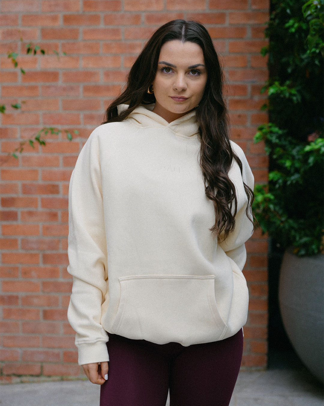 A woman wearing a cream-colored oversized BALI hoodie and dark purple BALI leggings, standing in front of a red brick wall with green plants.