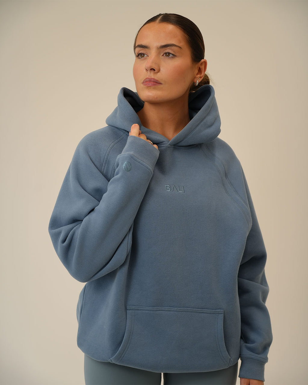 A woman wearing a dusty blue pullover BALI hoodie with a small tonal embroidered "BALI" logo on the chest, posed against a neutral beige background.