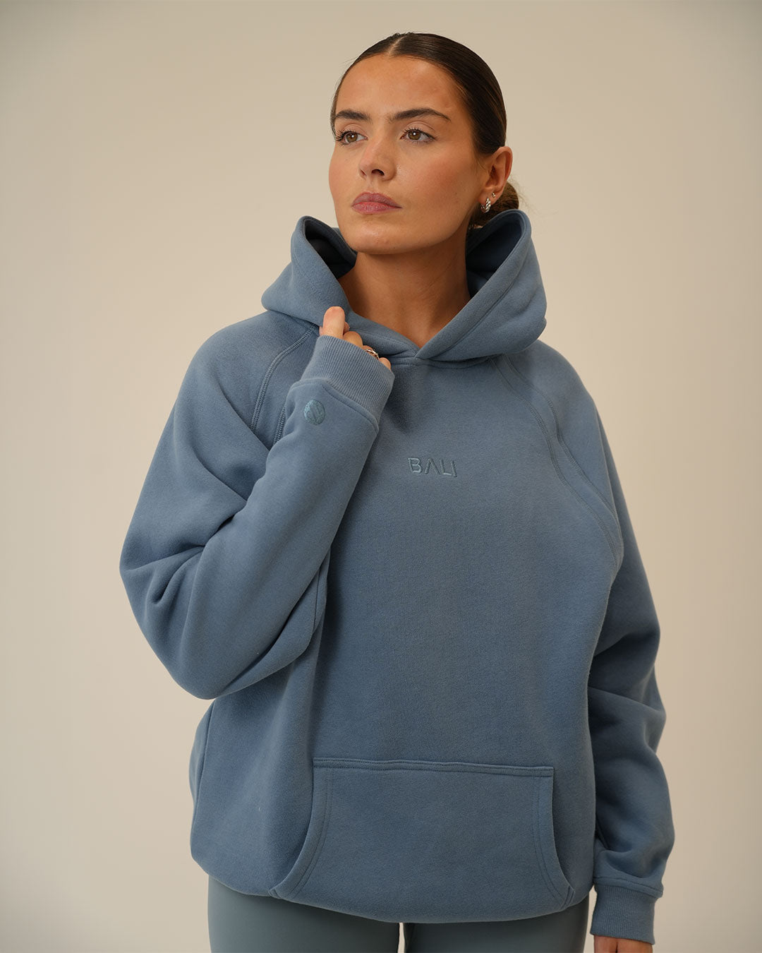 A woman wearing a dusty blue pullover BALI hoodie with a small tonal embroidered "BALI" logo on the chest, posed against a neutral beige background.