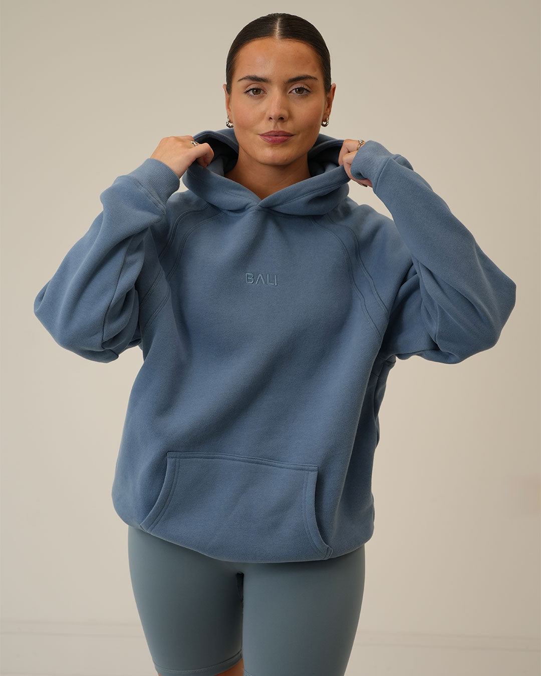 A woman wearing an oversized dusty blue BALI hoodie with a small tonal 'BALI' logo on the chest, paired with matching blue BALI shorts, against a plain beige studio background.