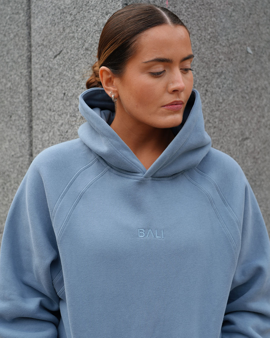 A woman wearing a light blue BALI hoodie with the brand name embroidered on the chest, standing against a gray textured wall.